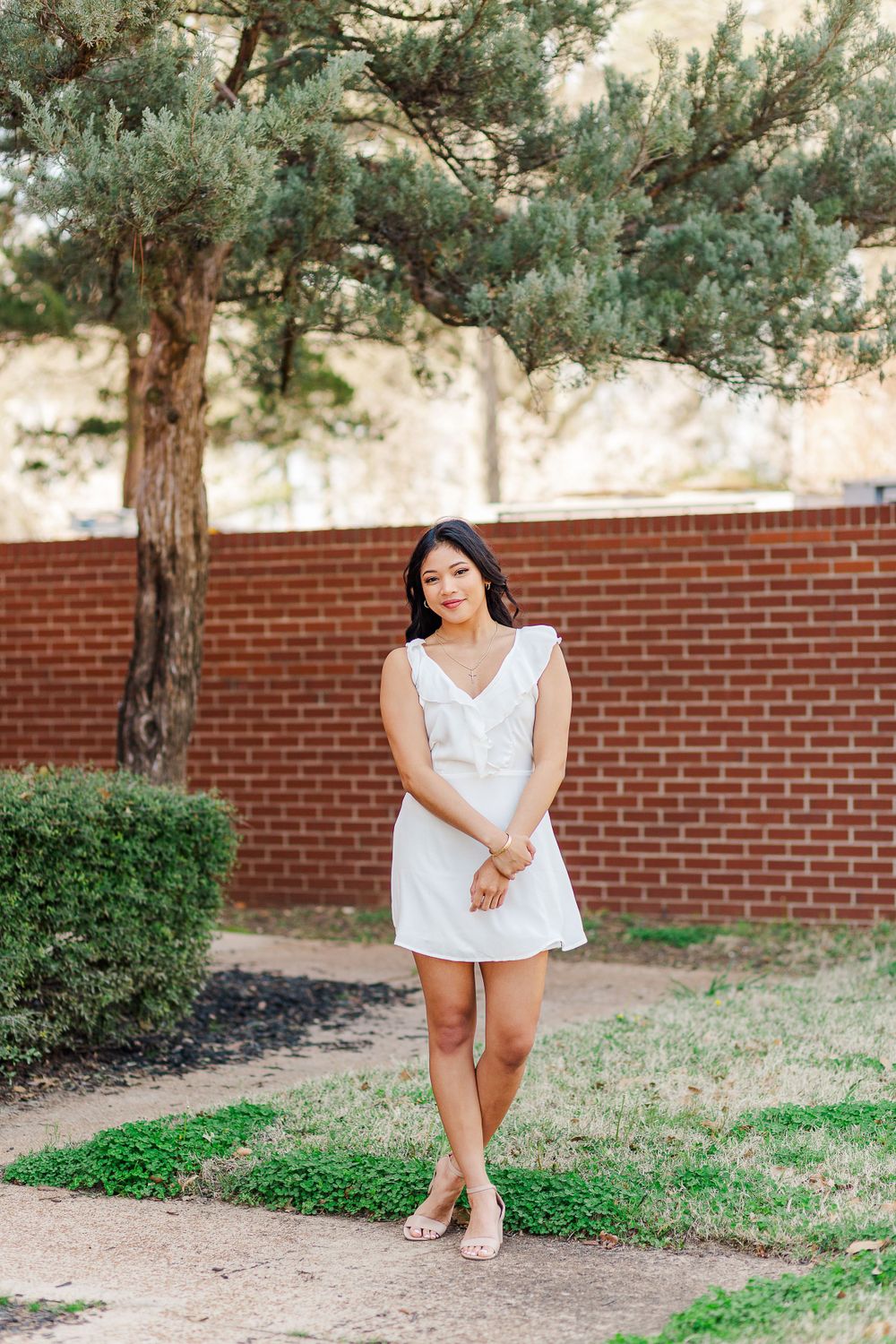 Woman in white dress poses gracefully on a path with pine trees and brick wall in background.