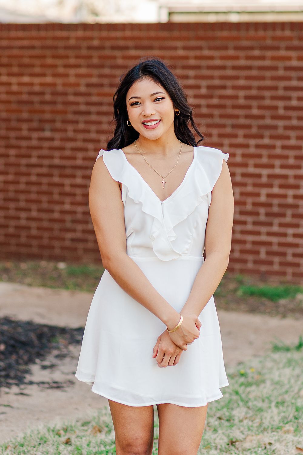 A graduate in white ruffled dress smiles during outdoor portrait session against brick backdrop.