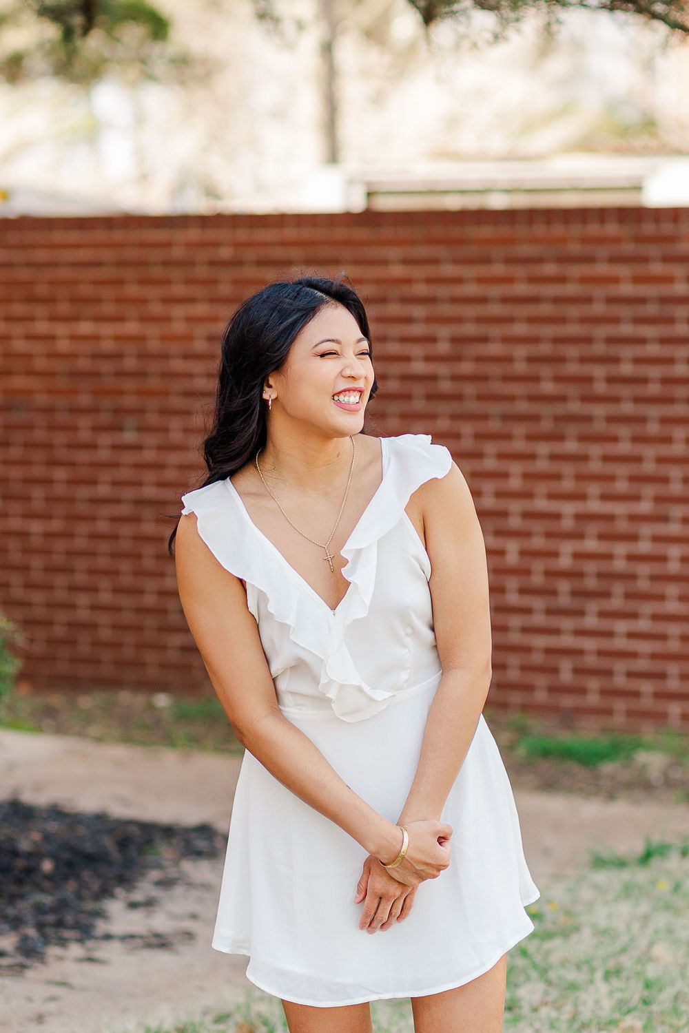 A joyful woman in a white ruffled sundress poses against a brick wall backdrop.