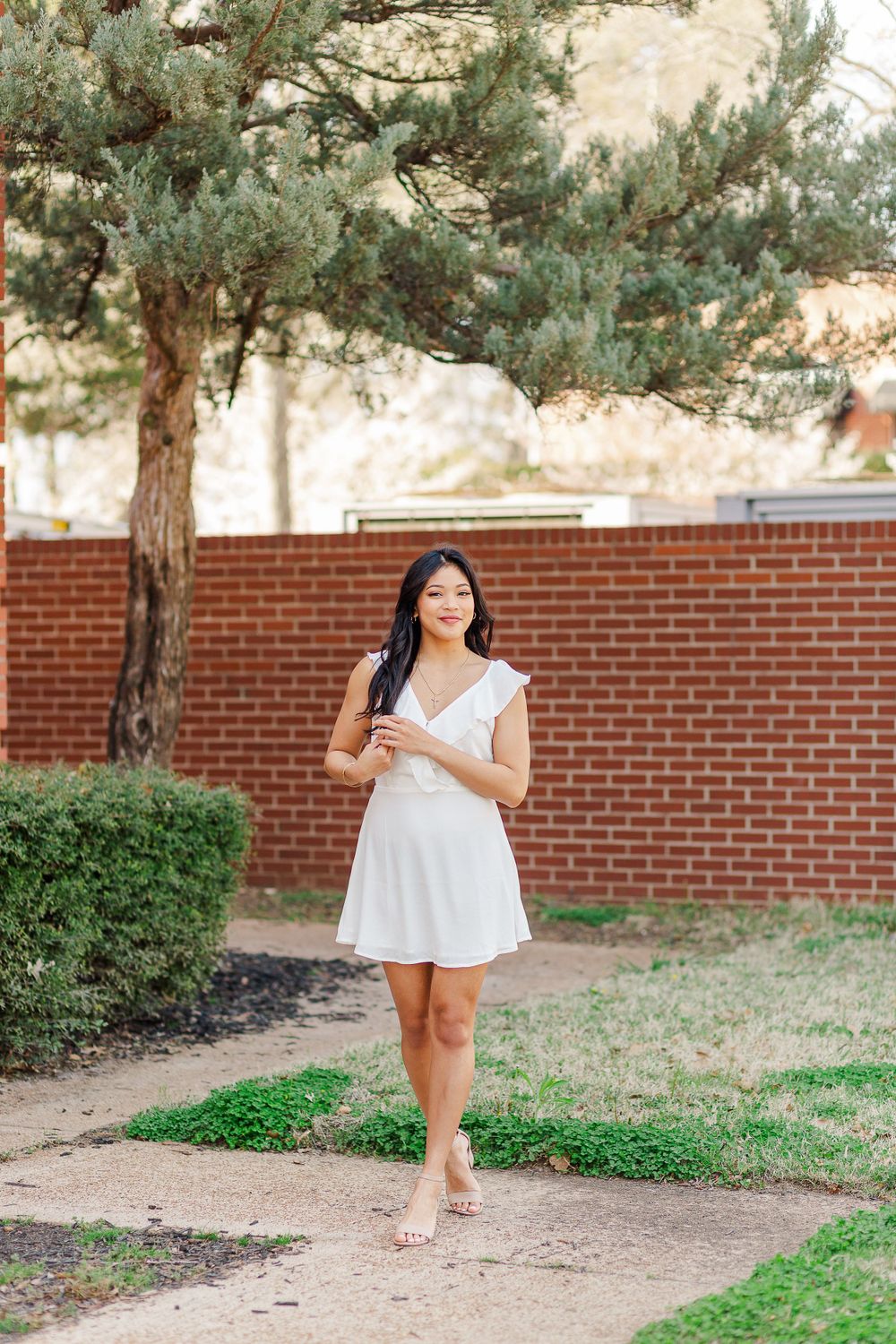 Woman in white dress strolls along garden path with pine trees overhead.