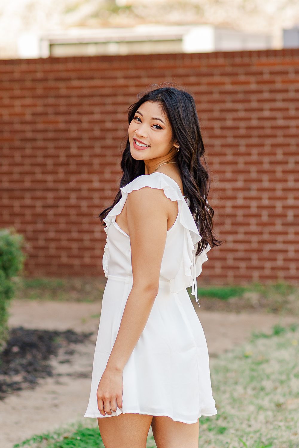 Woman in white ruffle dress poses playfully looking over shoulder near brick wall.