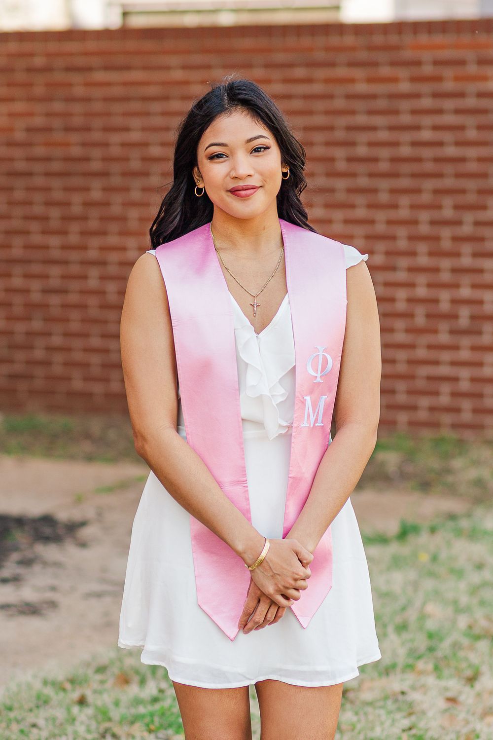Graduate wearing white dress and pink sorority stash poses against brick wall.