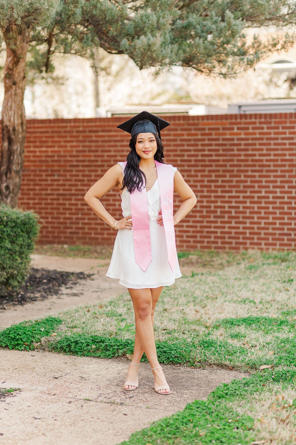 Graduate in white dress and pink stole stands confidently on garden path.