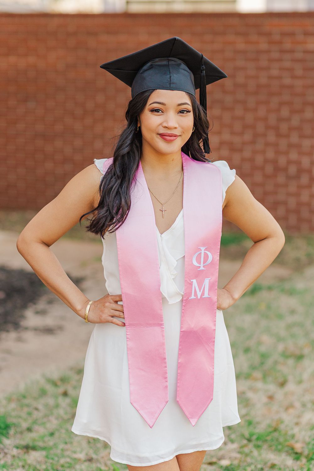 Graduate in white dress, pink stole and mortarboard cap poses for graduation photo.