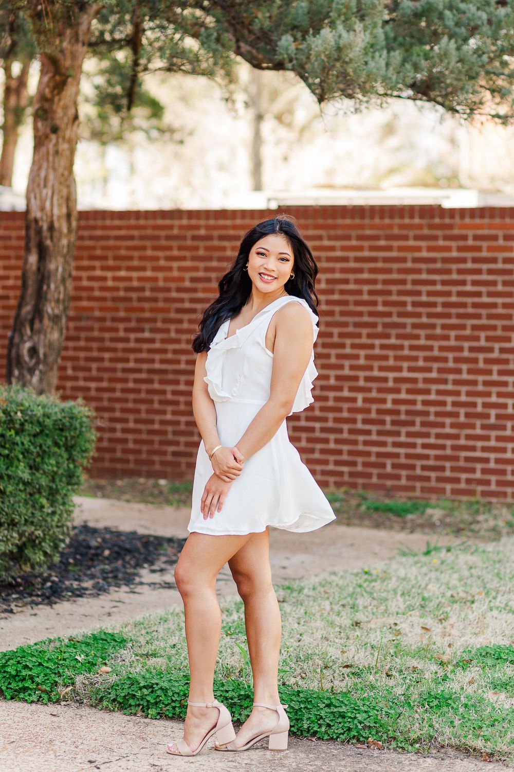 A graduate in white summer dress poses outdoors near trees and brick wall.