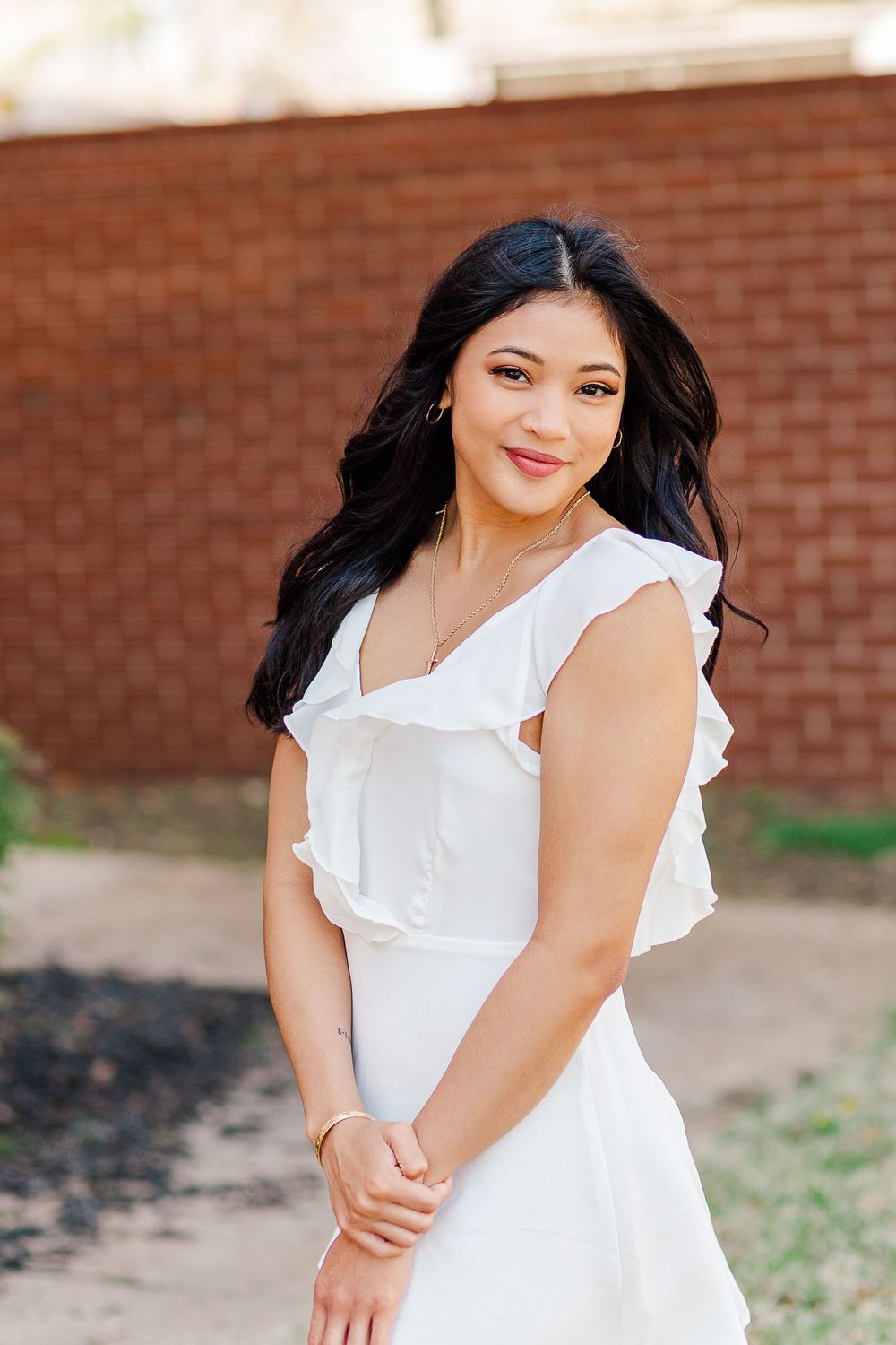 A graduate in a flowing white dress poses for portraits against brick wall background.