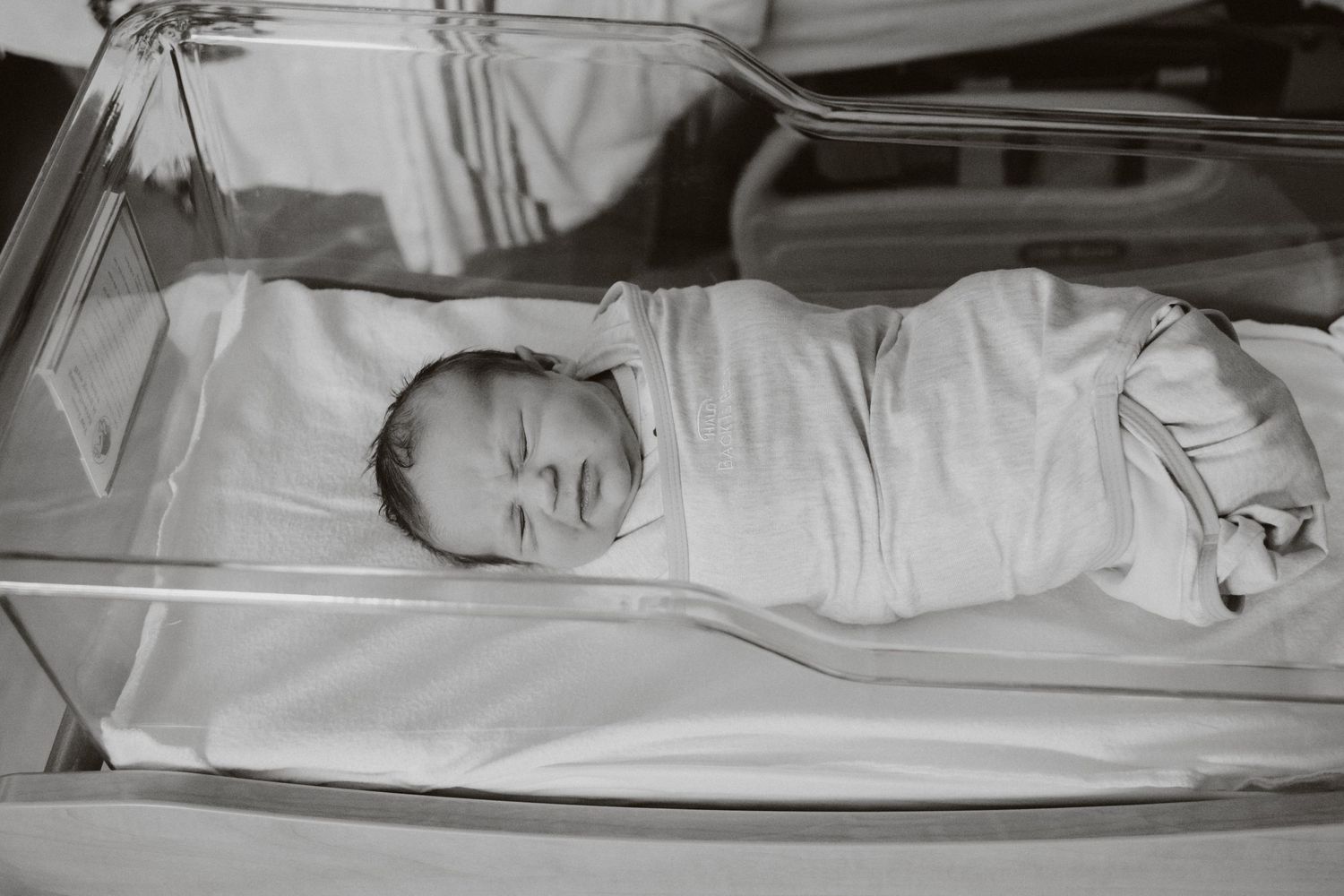 Black and white photo of sleeping newborn in hospital bassinet wrapped in white blanket.