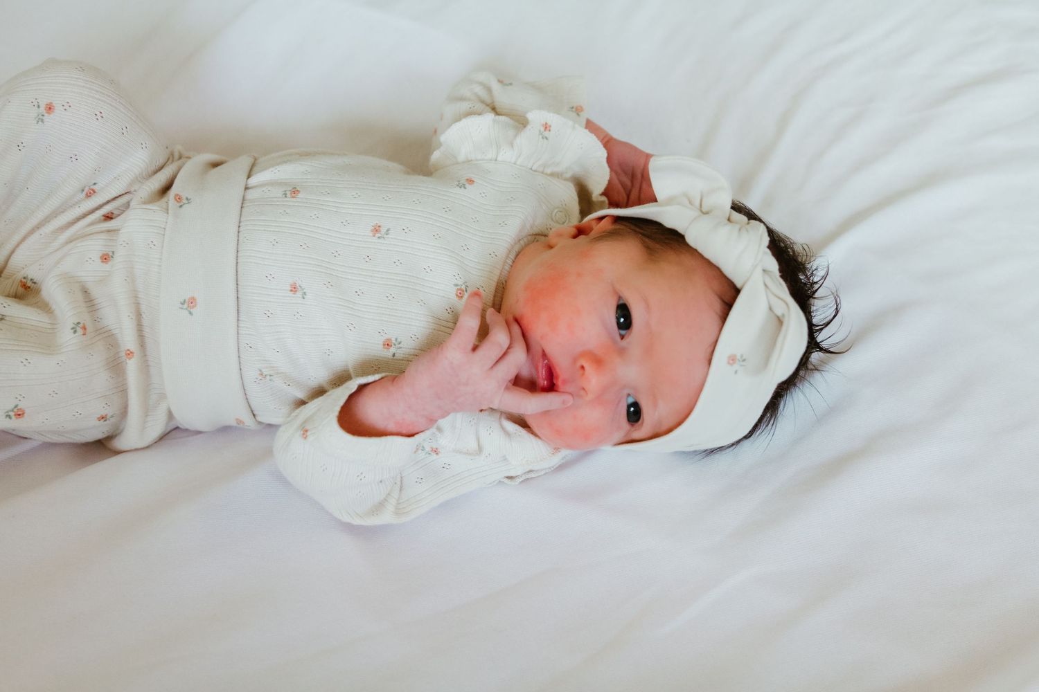A baby wearing a white outfit and headband lies on white bedding.