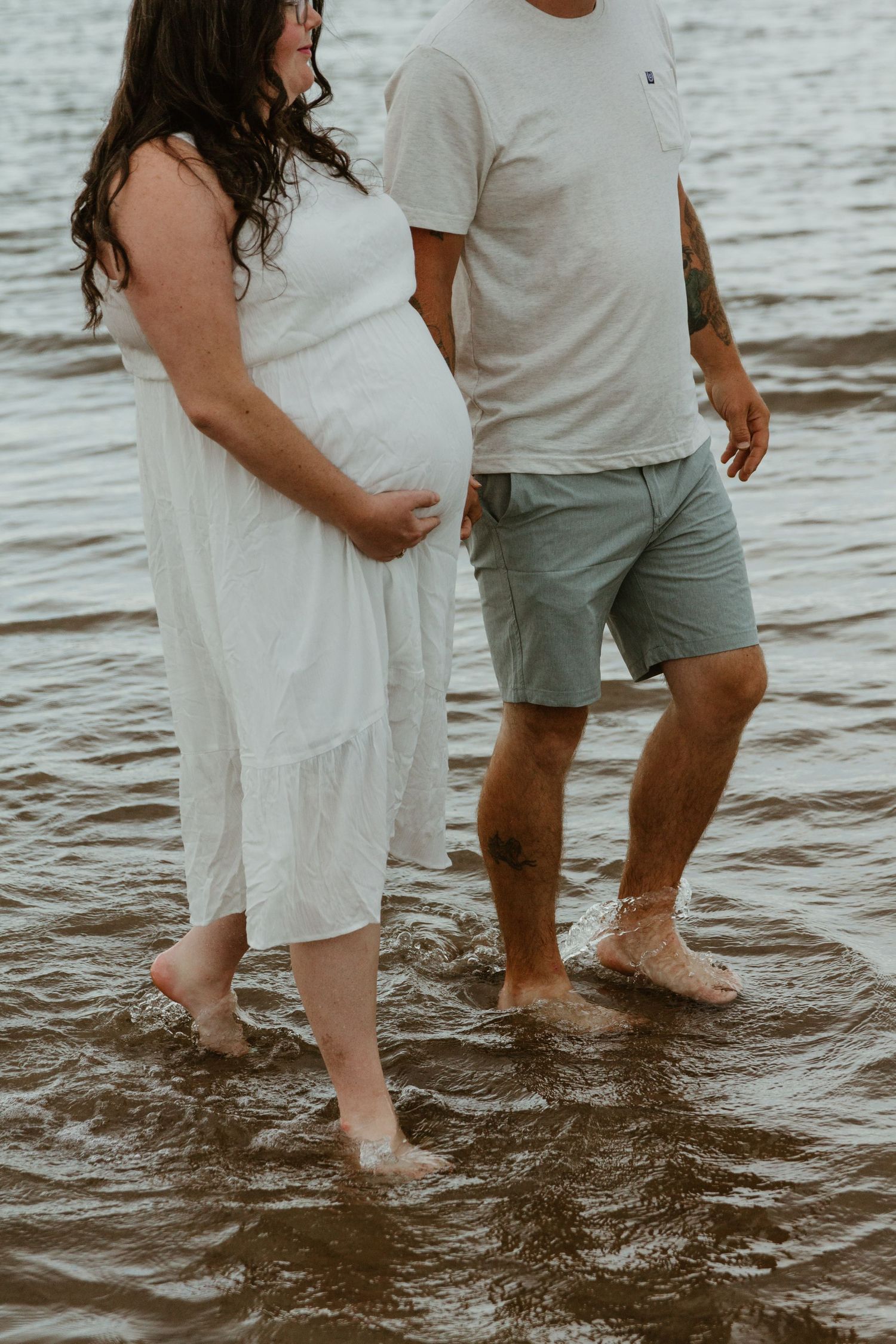 Expecting couple standing in shallow water at beach wearing white dress and casual attire.
