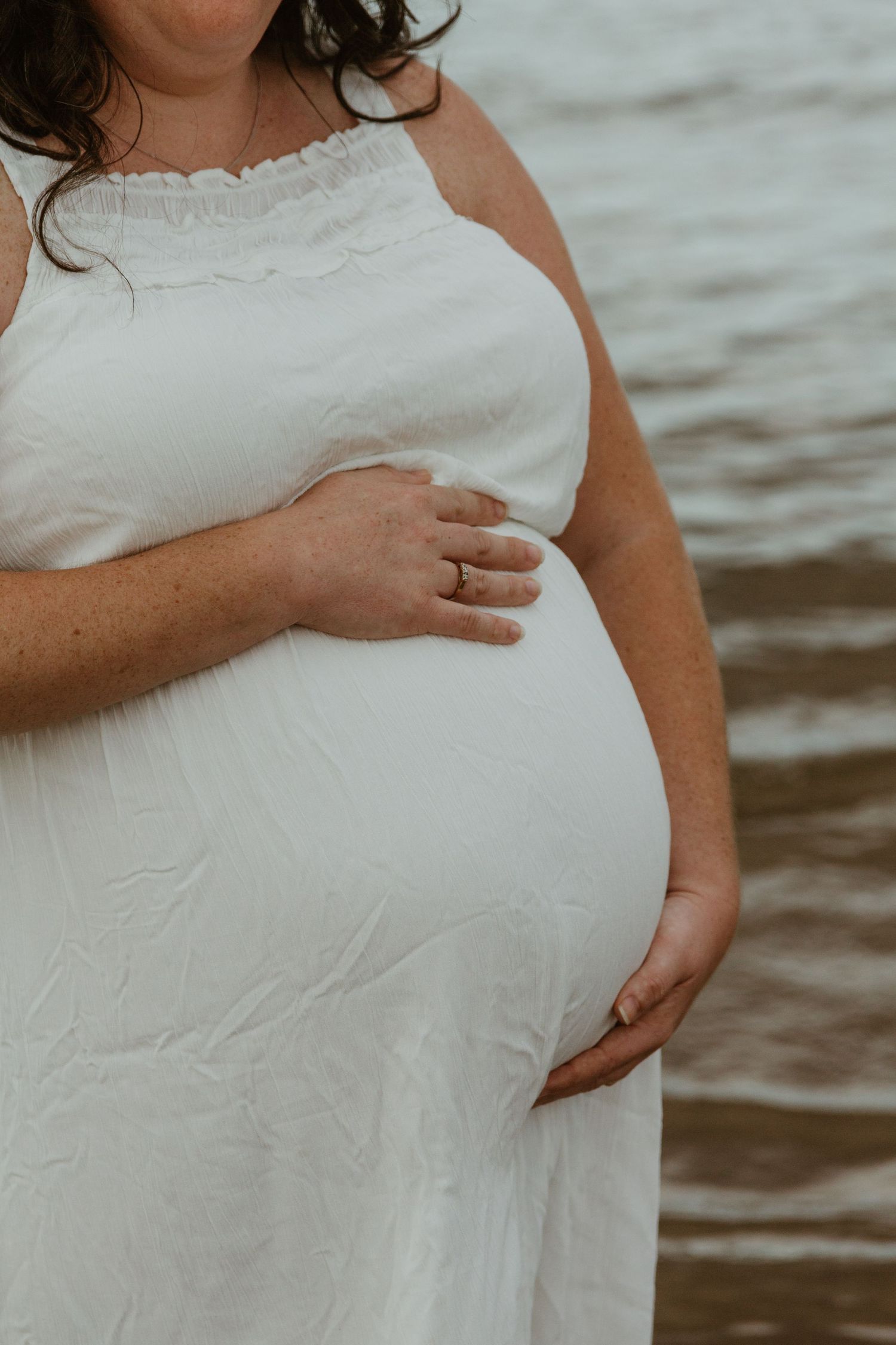 Close-up of pregnant woman in white dress standing by water showing baby bump.