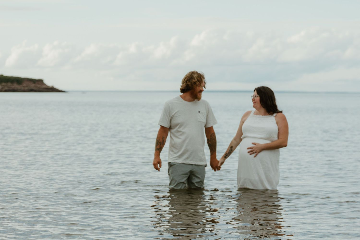 Couple holding hands while standing in ocean water against cloudy horizon.