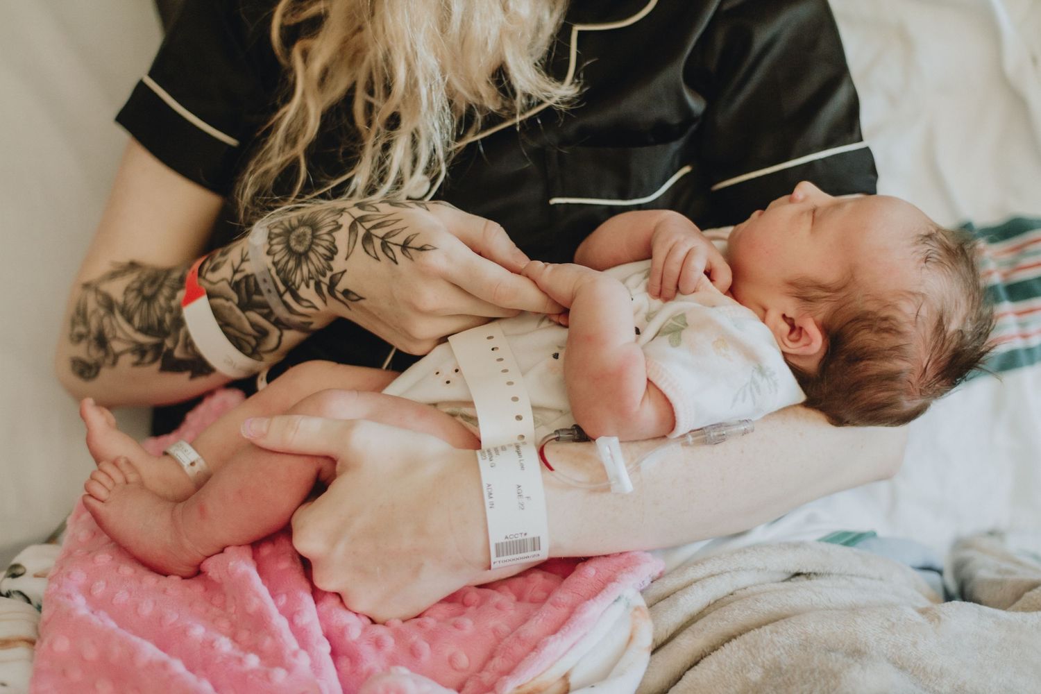 Parent with tattooed arms cradles newborn baby wrapped in white against pink blanket.