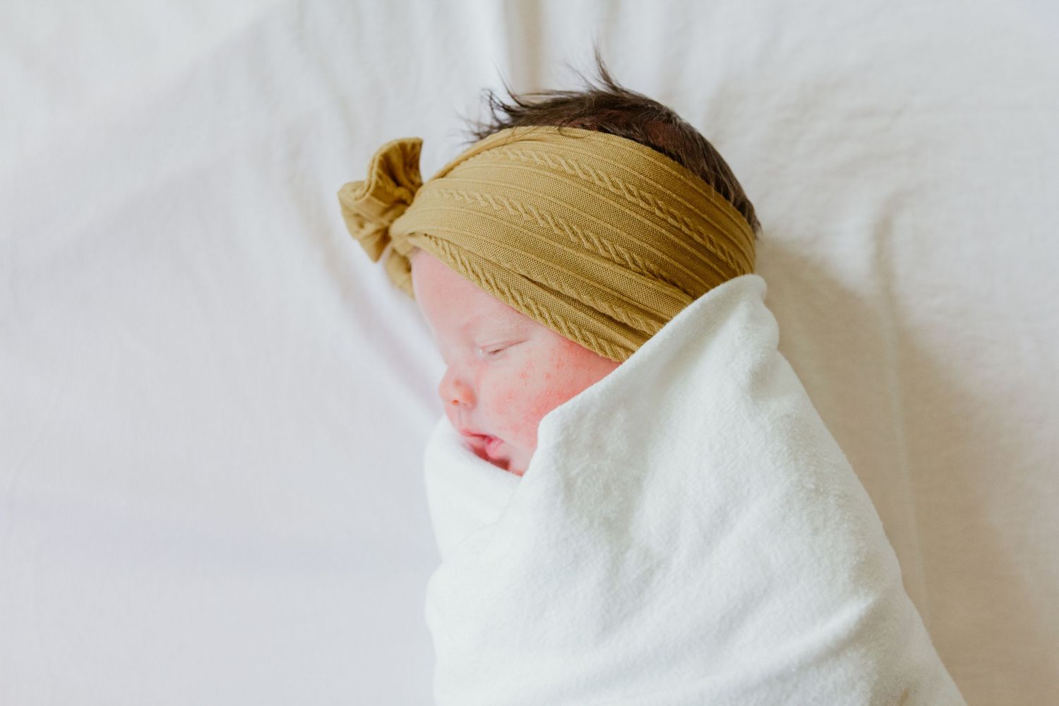 A sleeping newborn wrapped in white with a mustard yellow headband.