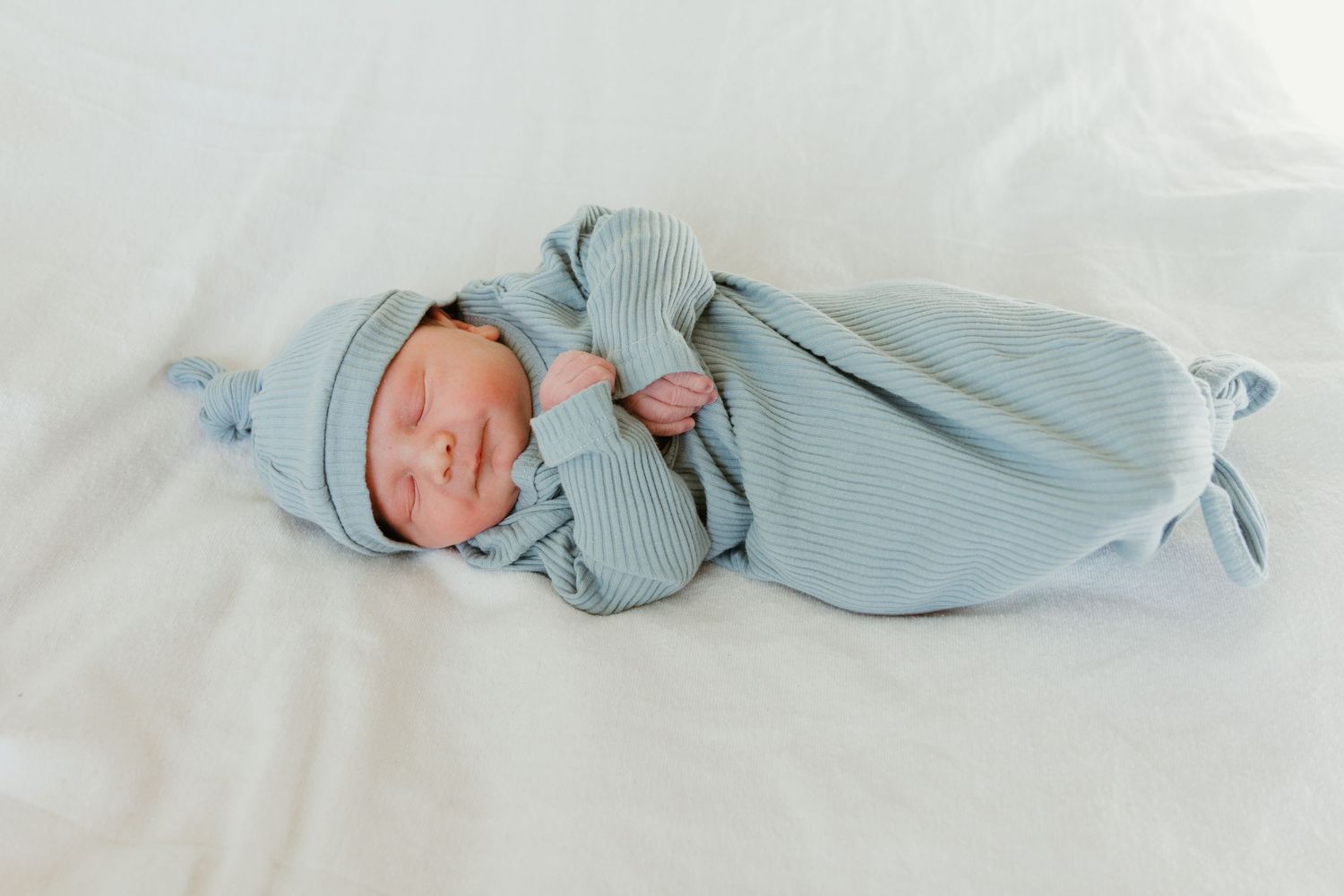 Sleeping newborn baby wearing soft blue swaddle and matching hat on white background.