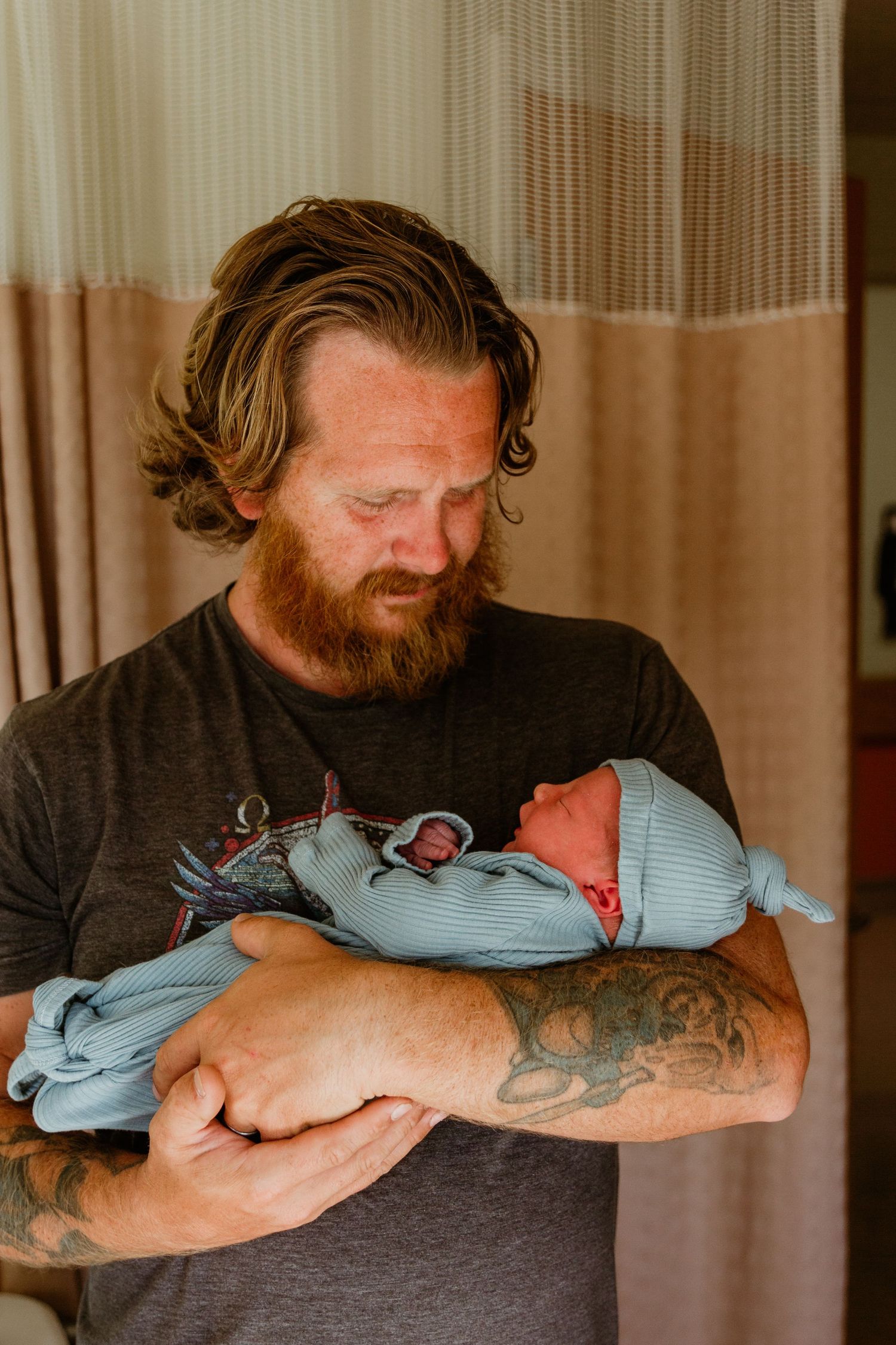 A bearded father in dark shirt tenderly holds his newborn baby wrapped in blue.