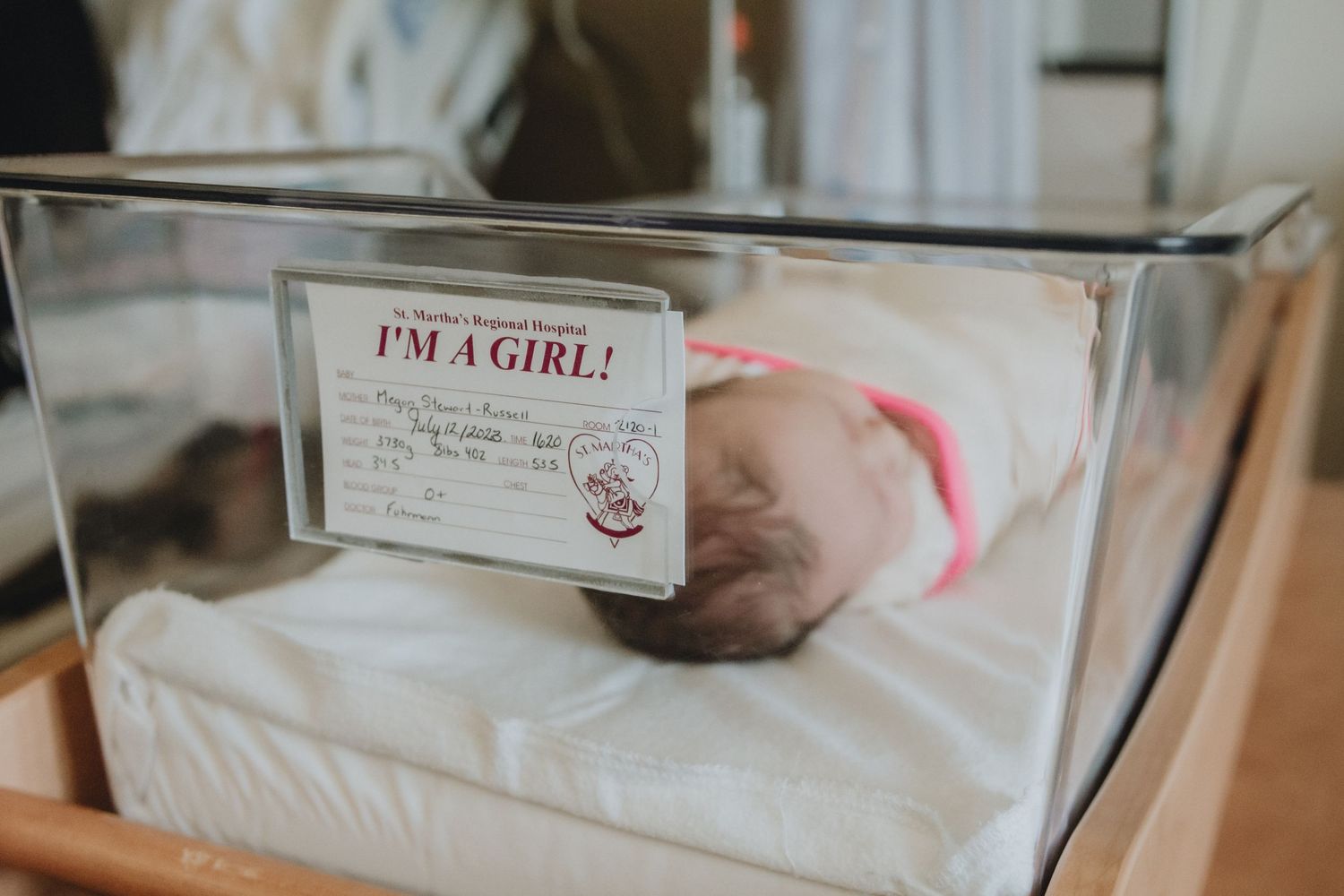 A newborn baby in a hospital bassinet with a 'I'm a Girl!' card displayed.