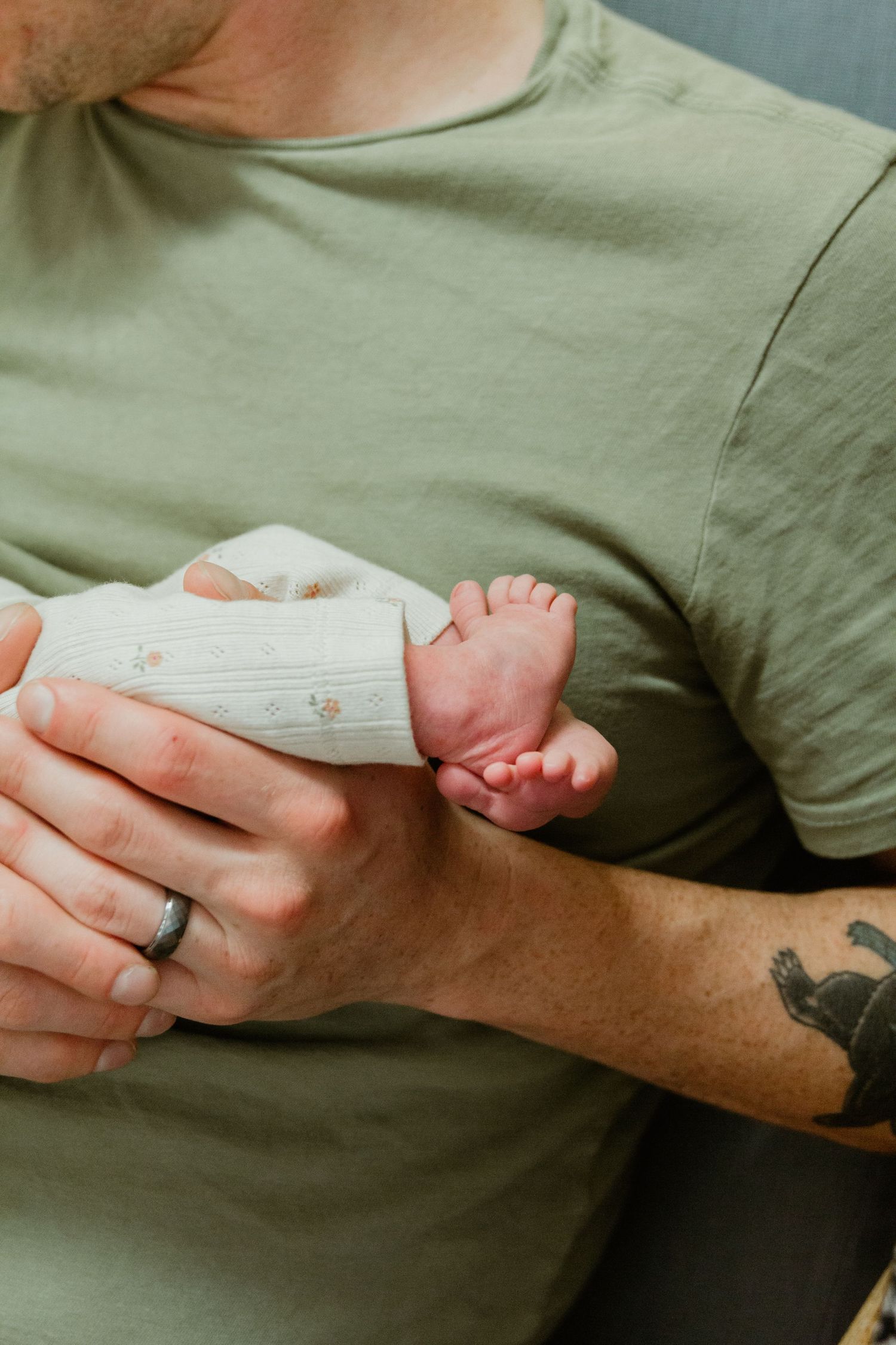 A close-up of small baby feet being held by hands with a wedding ring and arm tattoo.