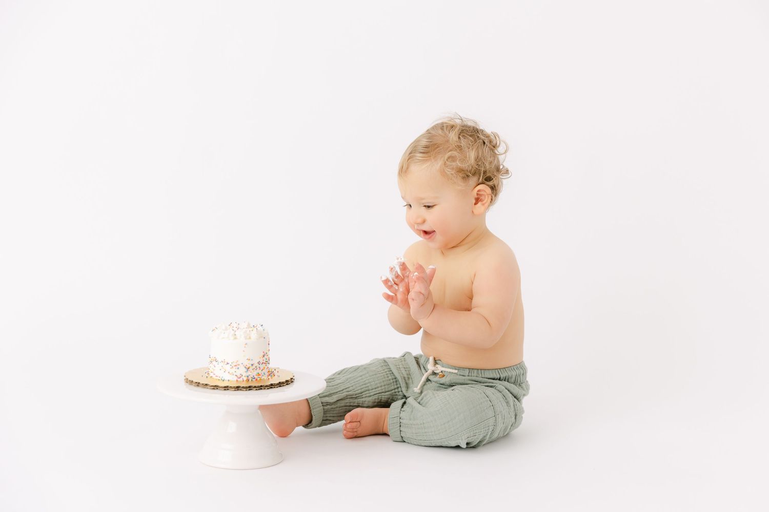 Baby with curly blonde hair wears light green pants and sits clapping his hands looking at a vanilla frosted cake.