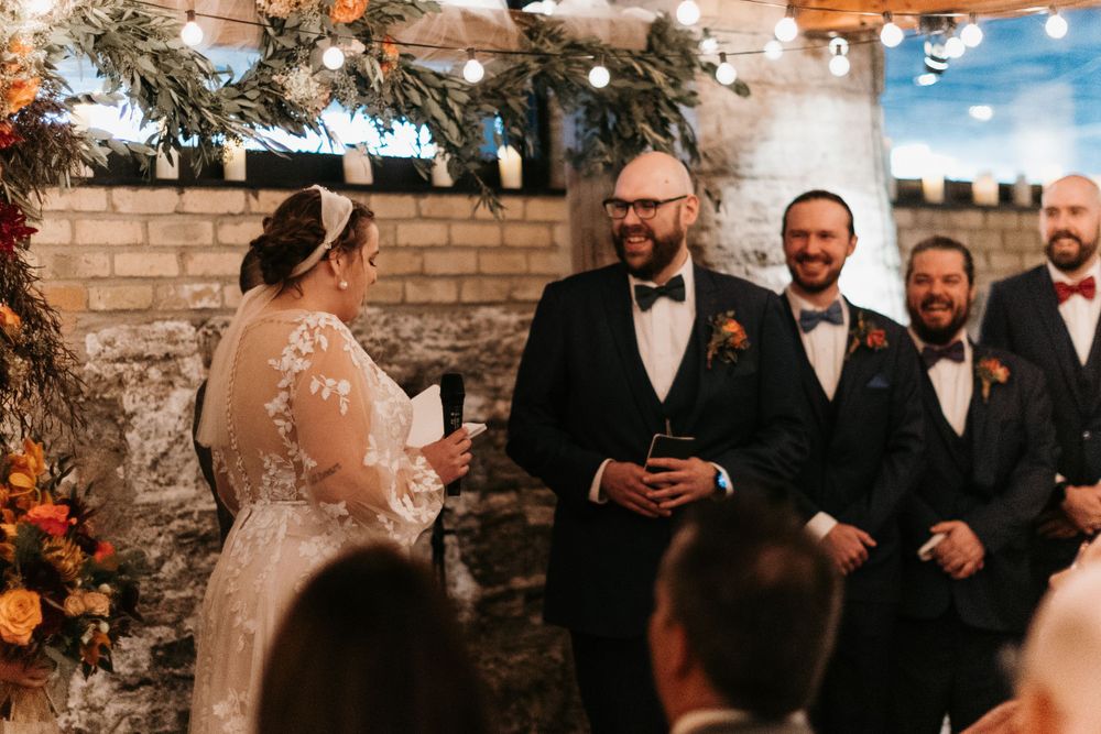 Groom in black tuxedo smiles at bride during indoor wedding ceremony with groomsmen and autumn floral decor.