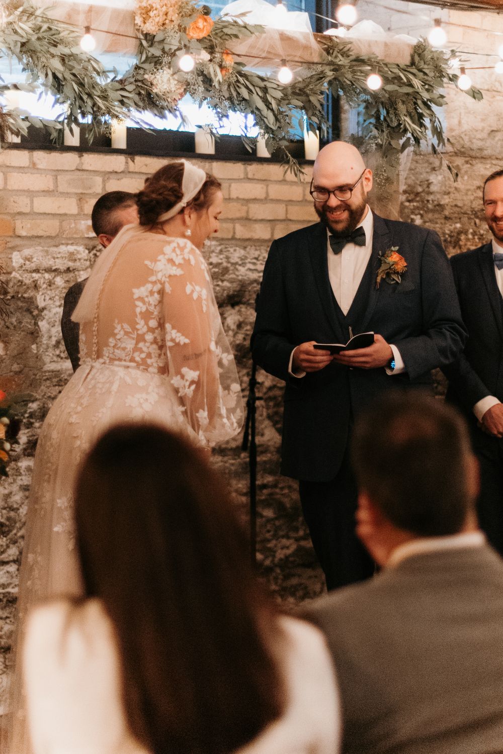 Groom reads vows from booklet as bride smiles at rustic indoor wedding ceremony.