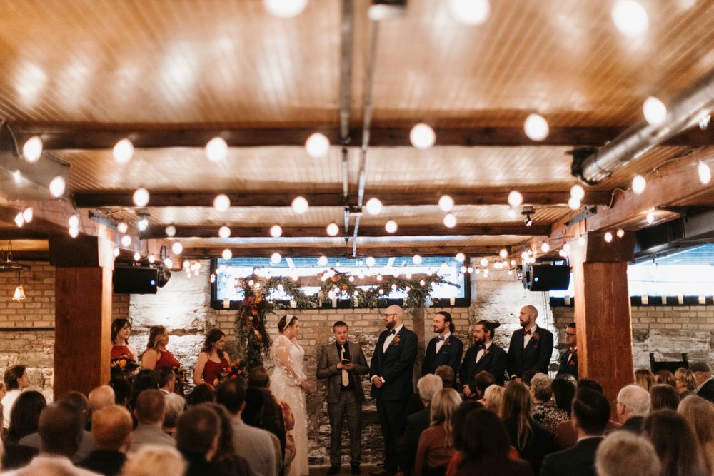 Elegant indoor wedding ceremony with string lights, wooden ceiling, and brick walls with wedding party at altar.