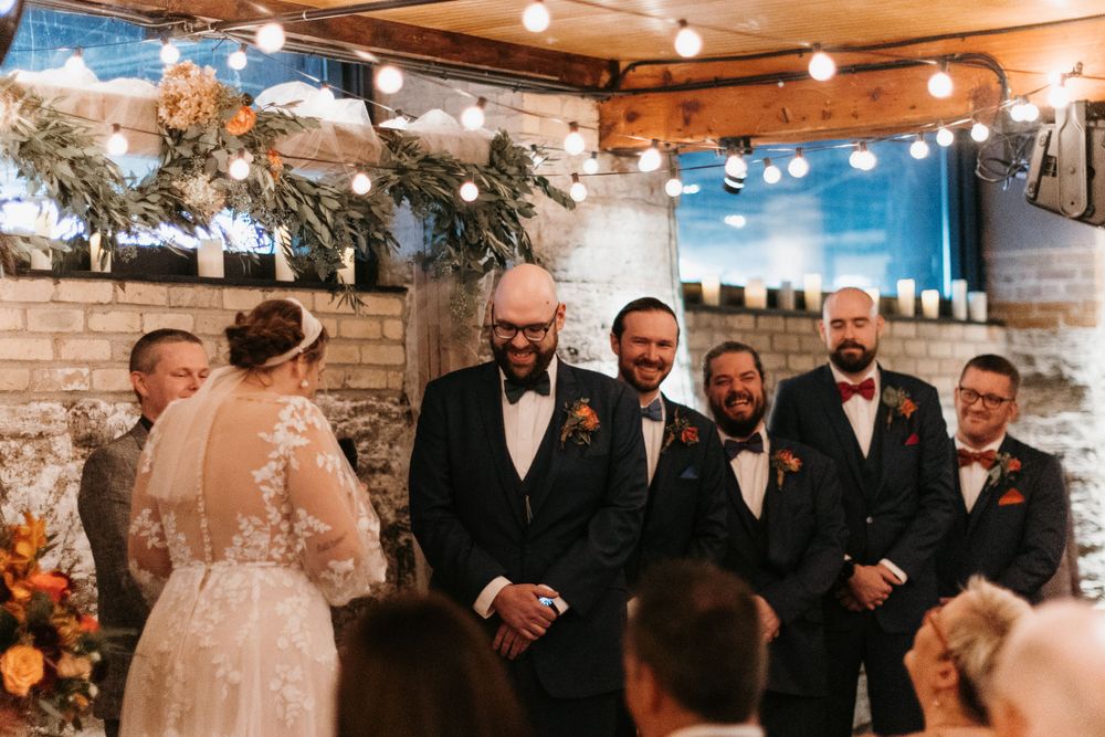 Groom smiles at bride during indoor wedding ceremony with groomsmen in navy suits.