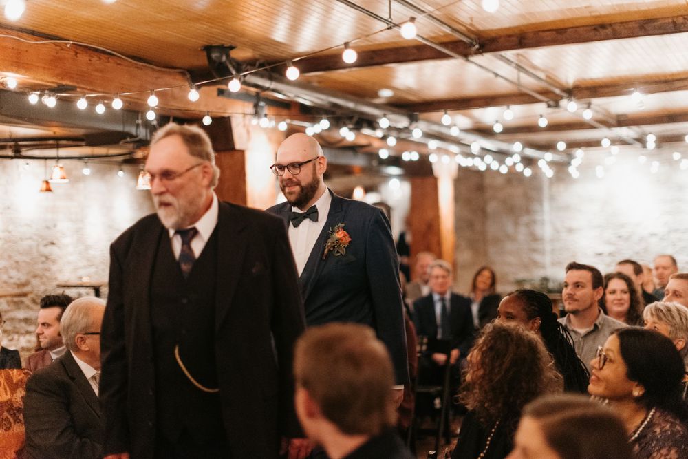 Groom and elder escort walk down the aisle at a rustic indoor wedding venue.