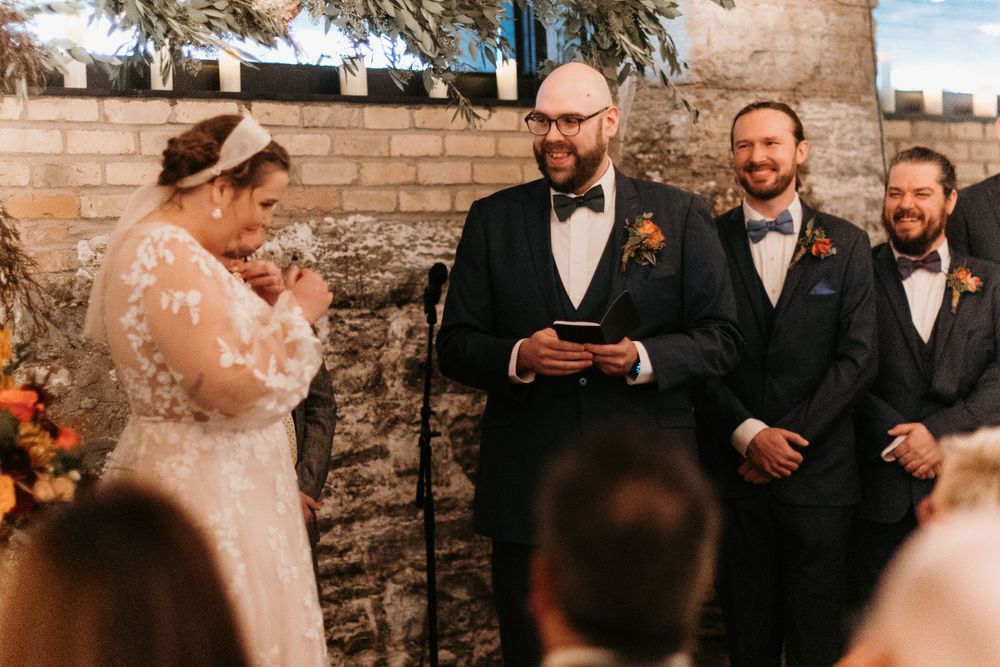 Groom reads vows as emotional bride wipes tears at rustic indoor ceremony.