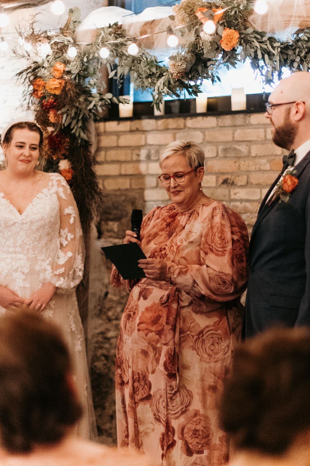 Wedding officiant in floral dress reads vows beside bride in lace gown and groom at brick wall altar.