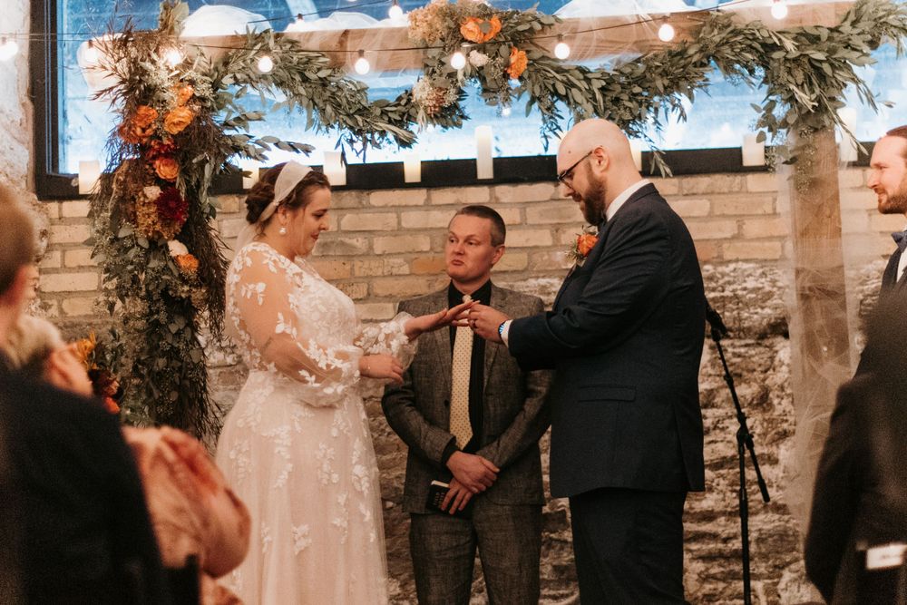 Groom reads vows to bride during wedding ceremony beneath lush greenery and fall floral arch indoors.