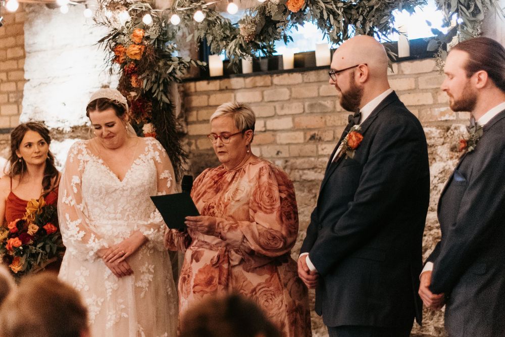 Bride smiles during indoor wedding ceremony with officiant and groom at autumn floral decorated brick wall altar.