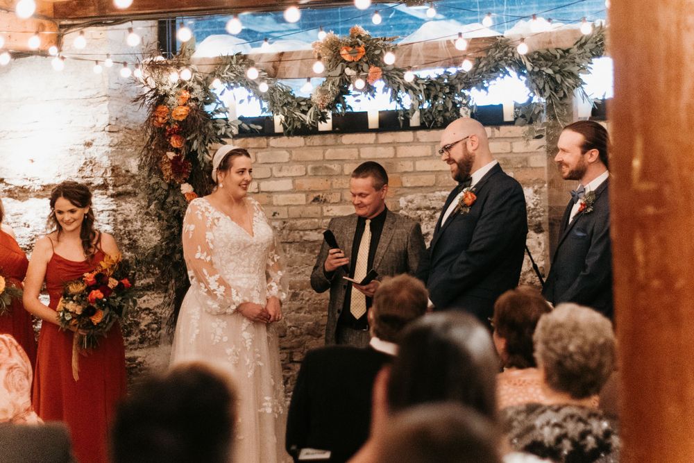 Bride and groom laugh during indoor wedding ceremony with officiant, string lights, and autumn floral arch backdrop.