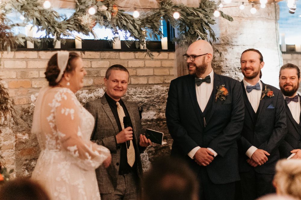 Groom in black tuxedo smiles at bride during indoor wedding ceremony with string lights and greenery backdrop.