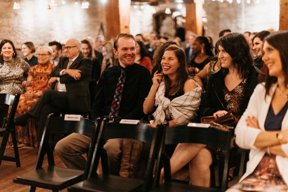 Wedding guests laughing and smiling while seated at an indoor ceremony.