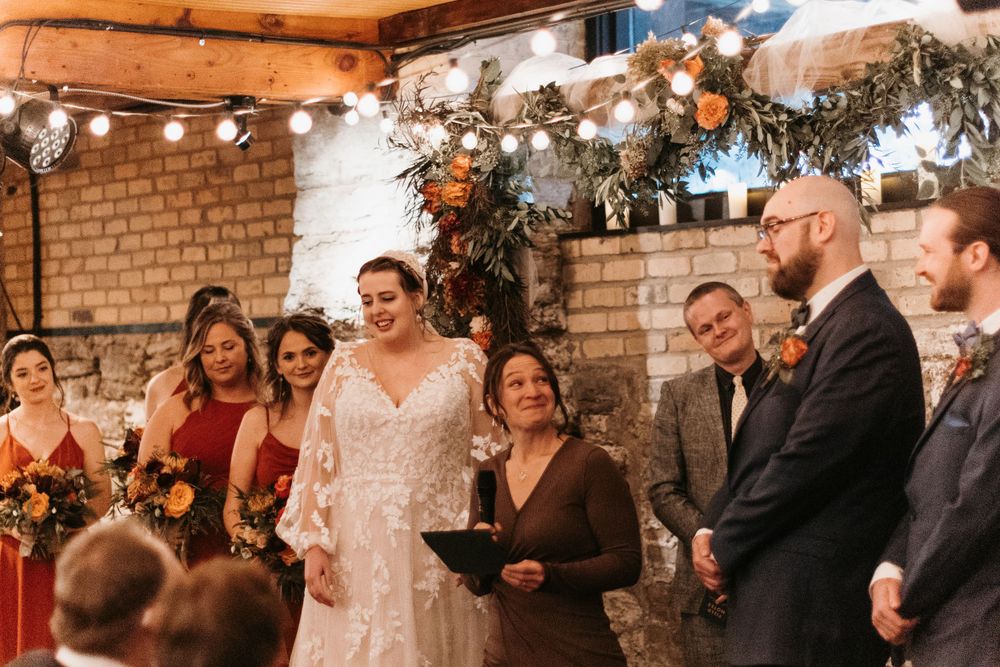 Bride and wedding party smile during indoor ceremony with officiant at autumn floral decorated brick wall altar.