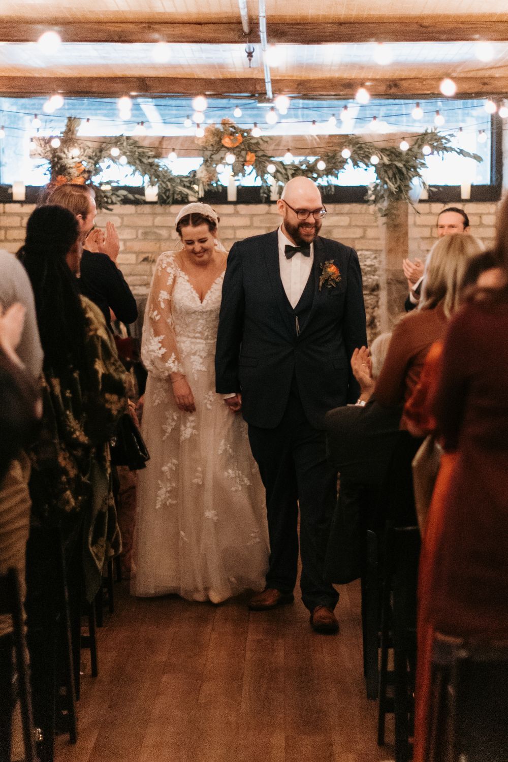Beaming newlyweds walk hand in hand down the aisle after wedding ceremony in rustic stone venue.