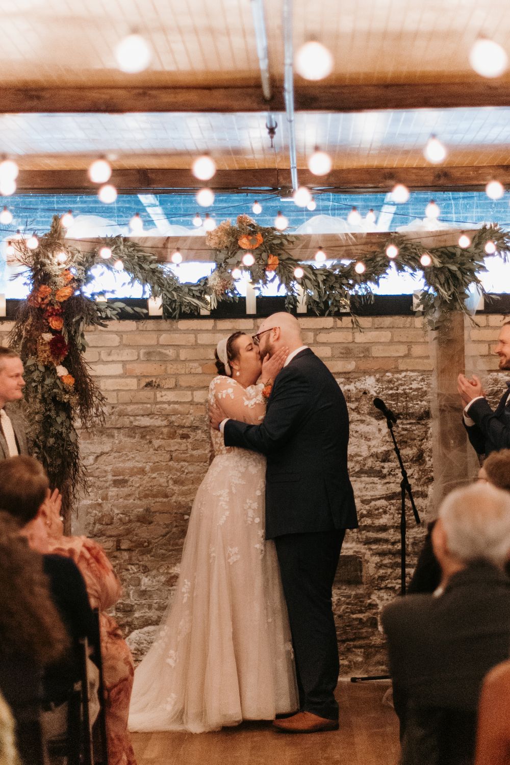 Bride and groom share romantic first kiss surrounded by applauding guests under Edison bulb string lights.
