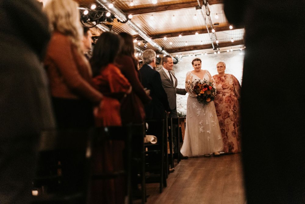 Bride walks down the aisle with escorts at a warmly lit rustic wedding venue.
