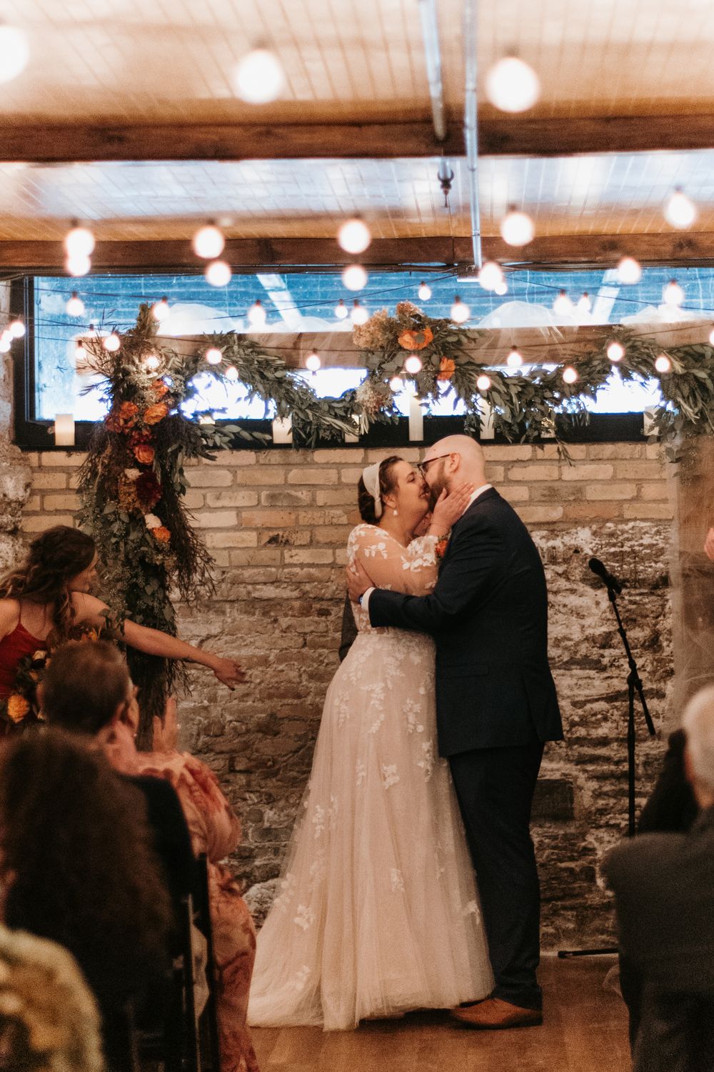 Newlyweds share their first kiss as a married couple under warm string lights at indoor wedding venue.