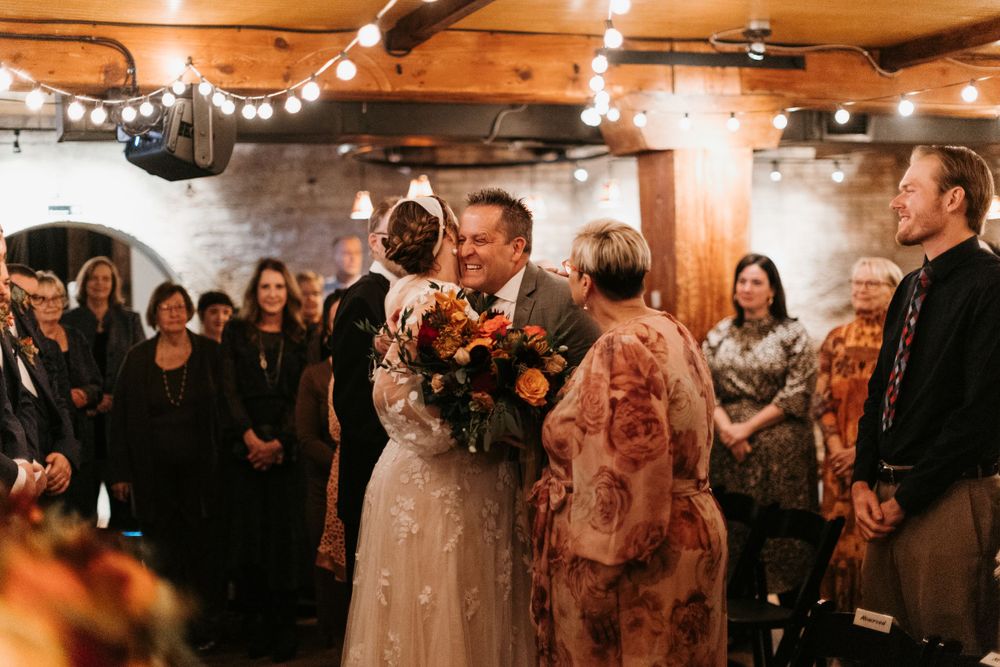 Bride and groom share first kiss at indoor wedding ceremony with string lights and autumn floral bouquet.