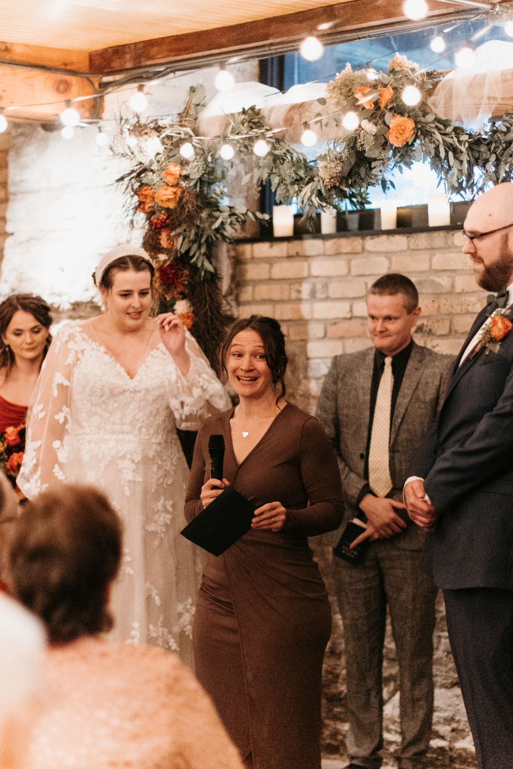 Emotional wedding officiant laughs as bride wipes tear during indoor ceremony at decorated brick wall altar.