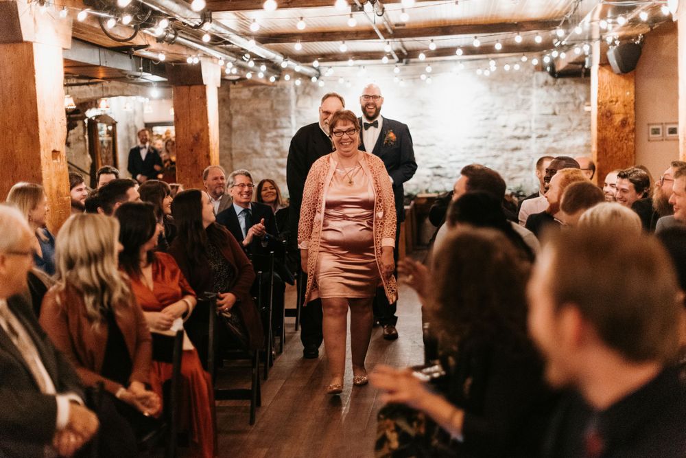 Mother of the groom walks down the aisle at a rustic indoor wedding ceremony.