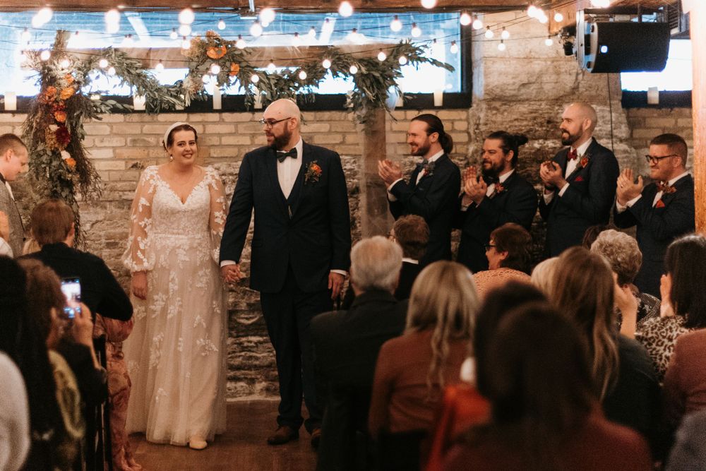 Joyful newlyweds walk back down the aisle hand in hand as guests celebrate at indoor wedding ceremony.