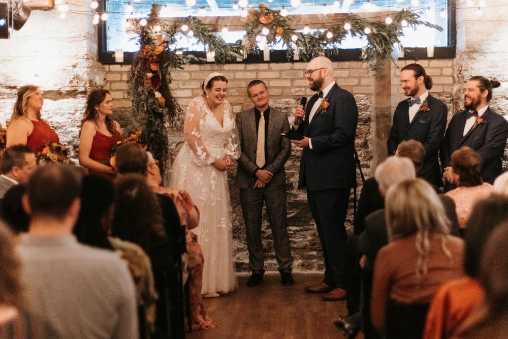 Couple exchanges vows at rustic indoor wedding with string lights and brick walls.