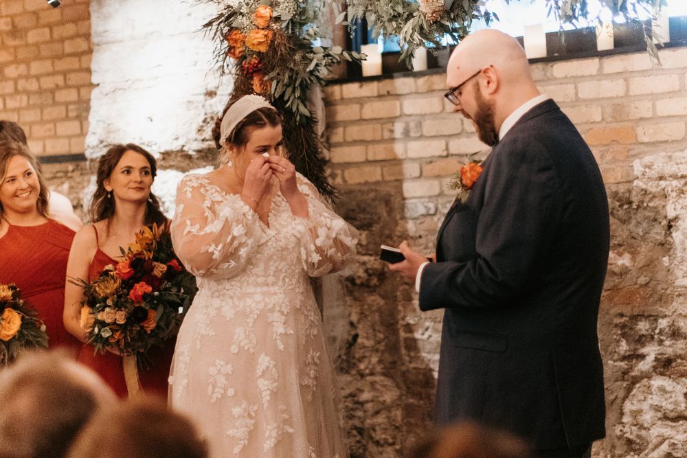 Emotional bride wipes tears as groom reads heartfelt vows at indoor ceremony.