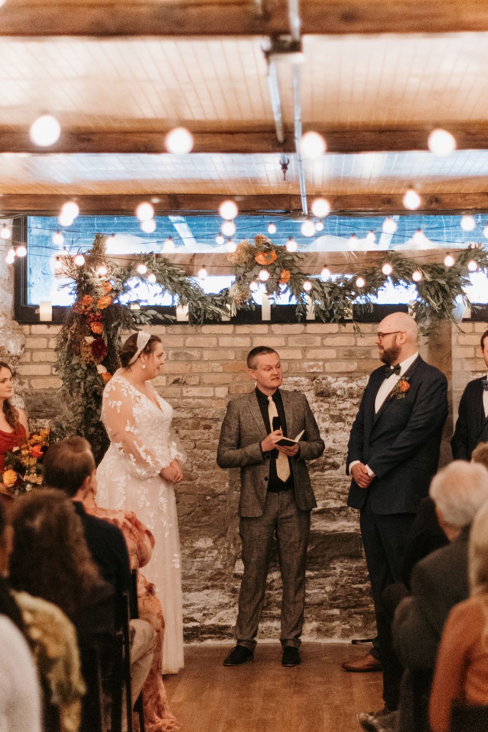 Bride and groom stand with officiant during indoor wedding ceremony beneath string lights and autumn floral arch.