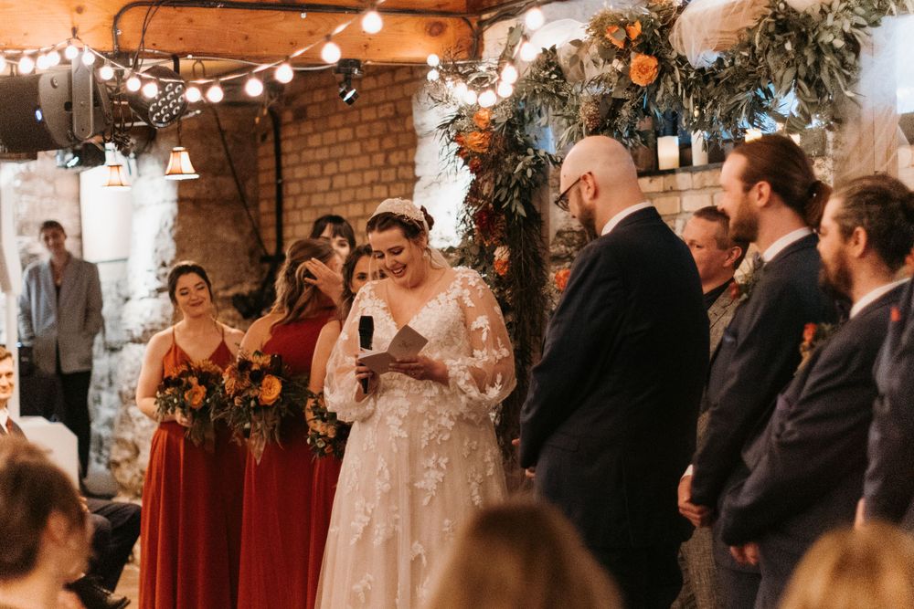 Bride reads vows to groom with bridesmaids in red dresses holding autumn bouquets.