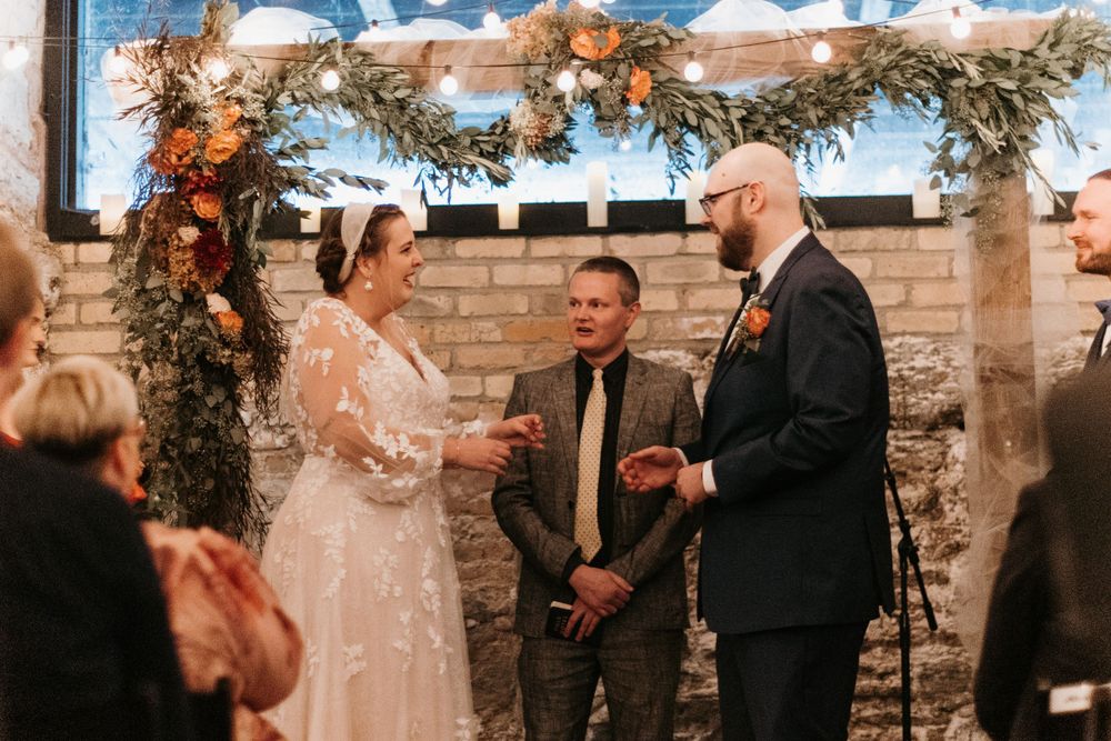 Smiling bride and groom exchange vows before officiant under a beautiful autumn floral arch with string lights.
