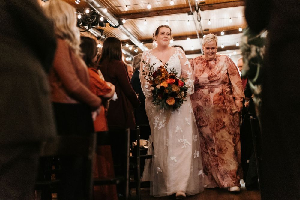 Bride in lace gown carries vibrant autumn bouquet while walking down the aisle.