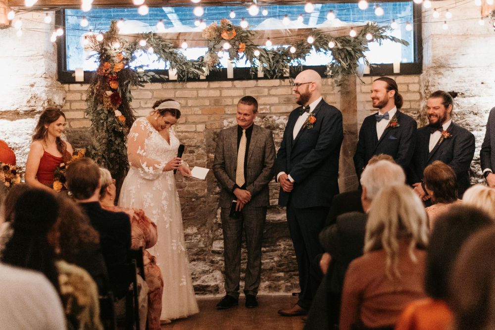 Bride reads vows at indoor wedding ceremony with string lights and floral backdrop.